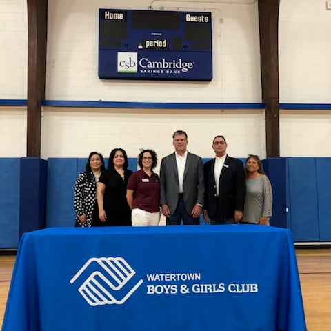 6 representatives from CSB or the Watertown Boys and Girls Club posing in a gym in front of a CSB digital scoreboard behind a table with the Watertown Boys & Girls Club logo on the tablecloth