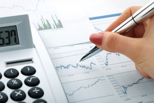 hand holding pen over paper with graphs with calculator beside it.