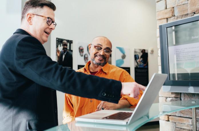 Man in black blazer and tortoise shell framed glasses points at a white laptop, indicating something on the screen to a bearded man in an orange button up shirt.