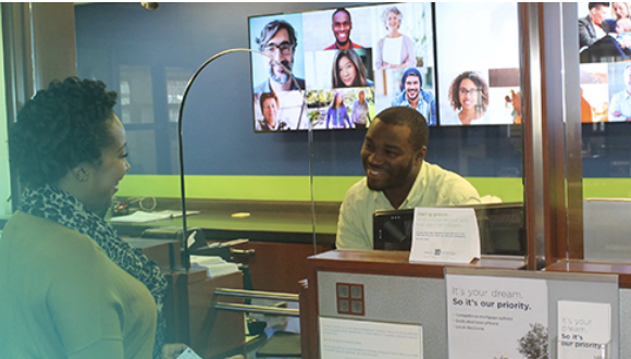 A male bank teller smiling and assisting a female customer from behind the glass