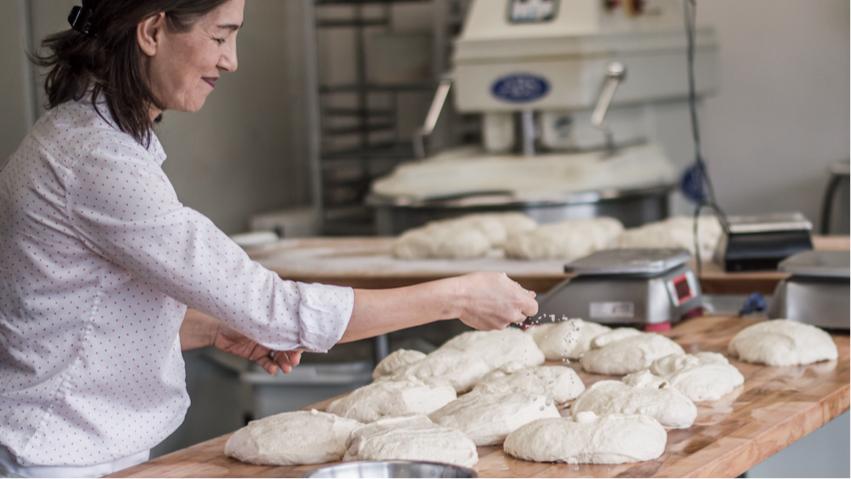 Small Business Bakery Owner with dark hair and white shirt prepares bread dough for rising by sprinkling salt on top.
