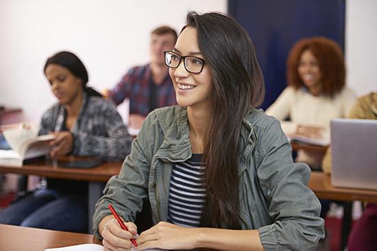Student in a classroom at desk with pen looking up smiling with 3 other students in the background