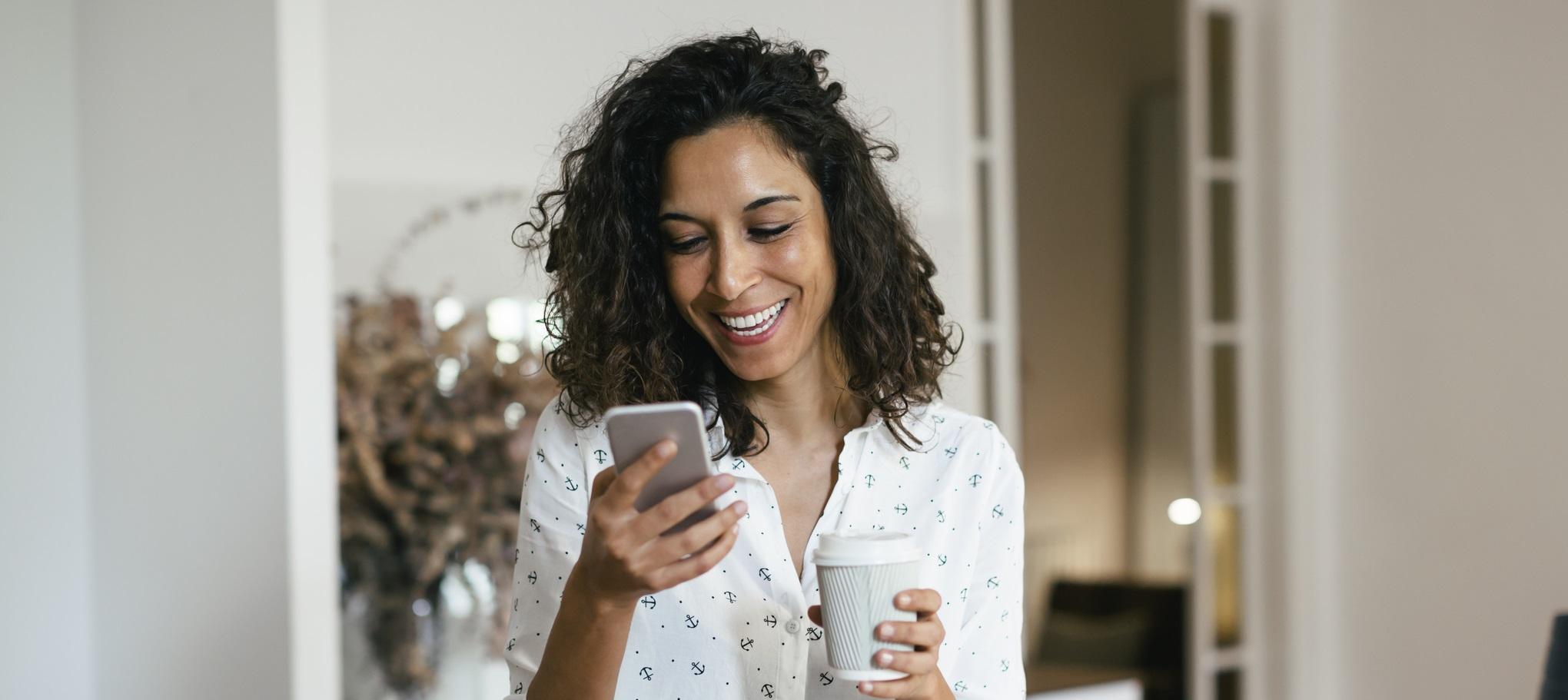 A woman holding a cell phone and a cup of coffee and smiling